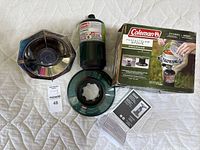 Full overhead view of the Coleman PerfectFlow stove set showing the stove base, propane canister, manual, and original box on a white quilted background.