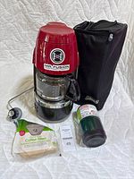 Red Coleman portable propane coffeemaker with glass pot, propane regulator, fuel canister, carrying case and coffee filters.