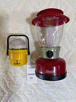 Photo of both battery lanterns, one large red Coleman lantern and one small yellow battery lantern side by side on white quilted fabric.