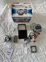 Photo showing the receiver box, receiver unit, remote control, wiring harness, mounting accessories, and instruction manuals arranged on white quilted surface.