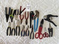 Full view of all 16 tools laid out on white quilted surface showing wide variety of pliers, pipe wrench, snips, scissors.