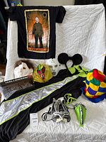 Photo of mixed lot costume accessories and masks arranged on white quilted backdrop. Items include multiple masks, a black 'The Devil' graphic T-shirt, colorful jester hat, costume robe/cloak, fuzzy ears headband, novelty rings, and frog hand puppet.