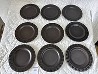 Nine black Pfaltzgraff stoneware dinner plates arranged in three rows on a white quilted surface, showing varied embossed patterns on edges and subtle differences in tone.
