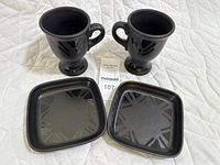 Two dark ceramic mugs with pedestal feet and geometric pattern next to two square dark ceramic plates/trays with rounded edges and same pattern