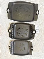 Top view showing three black rectangular Pfaltzgraff Midnight Sun stoneware trays with embossed starburst pattern, held handles at both ends.