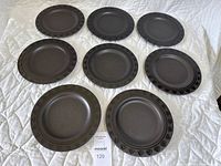 Top view of 8 black stoneware plates arranged on a white quilted surface, showing the front with decorative textured rim details.