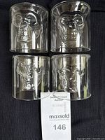 Front and side view of four clear glass whisky glasses arranged in two rows, each containing a raised three-dimensional skull shape inside.