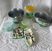 Front view of the entire lot showing 2 galvanized metal buckets with tropical pineapple design, 1 plastic drink pitcher with lid, 3 plastic containers with lids, oven mitt and pot holder with Hawaii Islands design placed on a white quilted surface