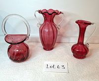 Front and side angle showing three cranberry glass items: basket with textured clear handle, vase with two clear handles, and jug with a single clear handle.
