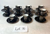 Twelve demitasse espresso cups and saucers arranged in two rows with bird and floral patterns on black exterior and gold trim, displayed with lot label.
