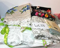 Group photo showing the folded floral embroidered runner, beige linen tablecloth with blue floral embroidery, and several white lace tablecloths tied with green ribbons with visible lace details and slight wear.