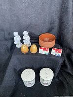 Photo showing all items arranged on black fabric background, including figurines, acorn salt and pepper, lobster trap salt and pepper, lobster-themed rectangular shakers, ceramic dish, and white ceramic shakers.