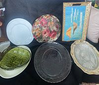 Front view of six assorted trays and bowls arranged on surface with white ceramic, floral metal, blue rectangular, green glass, silver-tone metal, and pressed glass items.
