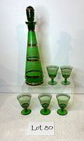 Photo showing the green glass liquor decanter with stopper and five matching stemmed glasses arranged around it. Gold banding visible, minor chip seen on one glass.
