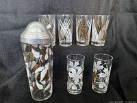 Cocktail shaker and six assorted vintage cocktail glasses with Mid-Century Modern patterns displayed on black fabric background.