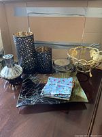 All 8 decorative home decor items displayed together on a table with cardboard background. Shows the paired black and gold candle holders, clear ribbed glass vase, metal lidded container with round coasters inside, gold leaf-shaped decorative bowl, square decorative plate with modern abstract pattern, bundle of blue cloth napkins with red bird print, black square decorative tray beneath.