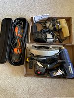 Top-down image showing assorted power tools including angle grinder in its case, drills, jigsaw, and heat gun arranged in two cardboard boxes.
