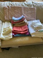 Image of various folded tablecloths and napkins in assorted colors and fabric blends stacked on a beige sofa.