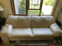 Front view of beige leather sofa placed in front of window with stained glass panel. Shows two seat cushions and padded armrests in natural light to highlight texture and creasing of leather upholstery.