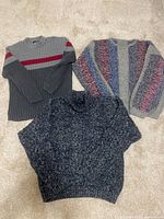 Three men’s sweaters laid out on carpet floor. Left is a gray ribbed knit with red stripe, middle is a dark blue and black marled knit, right is a multicolored striped textured knit sweater.