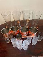 Clear tall glasses lined up behind copper Moscow Mule mugs with two glass swizzle sticks standing inside mugs, textured glass shot glasses and patterned tumblers in front.