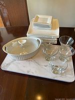 Mixed dinnerware lot with square ceramic plates, ceramic covered casserole dish, clear glass bowls and glasses, and marble-patterned serving tray