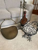 Brass fireplace tool set including stand and four tools, next to a decorative metal basket with twig design sitting on carpet.