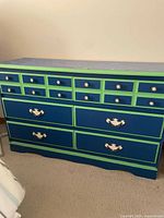 Wooden dresser painted in green and blue with 12 small drawers at the top with round metal knobs and 4 large drawers below with decorative metal handles.