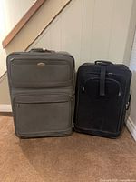 Photo shows two upright suitcases against a wall under stairs. One large gray suitcase with wheels and front zippered pocket, one medium black suitcase with front straps.