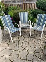 Four stackable outdoor patio chairs with white metal frames and blue-green striped fabric sling seats arranged outside on stone patio.
