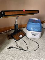 Photo showing a vintage gooseneck desk lamp with faux wood trim and a cool mist humidifier with a translucent blue tank. Both devices are on a carpeted floor, plugged in with corded power supply, with no visible damage or wear.