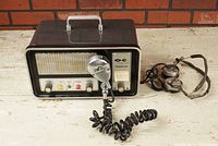 Front view of Hammarlund Outercom FM50A VHF-FM tube radio with microphone connected and headphones placed to the side on white wooden surface against brick wall background.