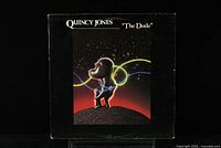 Front cover of the vinyl record album 'The Dude' by Quincy Jones. Artwork shows a dark figure holding brightly colored lines on a cosmic background.