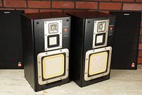 Pair of Sony APM-910AV 3-way speakers shown frontally with their grills placed behind, displaying the cloth surround drivers and the layout of tweeter, mid-range, and woofer.