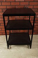 Front view of black metal frame rack with three wood-patterned shelves against a brick wall background.