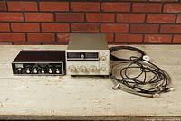 Wide view of both antenna tuners, Yaesu FC-107 in cream metal case, MFI Versa Tuner II black case, with cables coiled beside them on a wooden surface