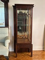 Front view of antique curio display cabinet showing wood carvings, glass door with gothic arch design, and mirrored back inside.