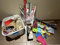 Photo showing plastic bin filled with assorted Christmas tree decorations including gold and red ornaments, tissue paper, and ribbons, with rolls of Christmas-themed wrapping paper and colorful gift bags arranged on the floor beside it.