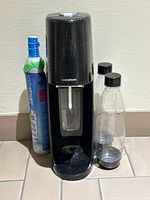 Front view of black SodaStream machine with two bottles and CO2 canister on floor against wall.