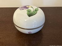 Top and side views of the white porcelain jewelry bowl showing hand-painted purple, yellow flowers and green leaves with gold accent band around the lid.