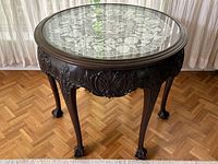 Full view of ornate dark wood round accent table showing claw feet, carved details on apron and legs, glass top, and lace doily underneath.