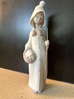 Full view of the Lladro figurine showing the girl holding a basket and a staff, in a white hooded coat standing upright.