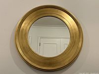 Front view showing the round gold leaf frame and reflective mirror surface, hung on a wall with visible door reflection.