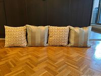 Four throw pillows arranged in a row on a wooden parquet floor against a dark wall, showing one side clear enough to see the damask and striped patterns and some wear/staining.