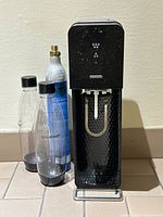 Black SodaStream machine with two clear plastic bottles and one CO2 canister in the background against a cream wall.