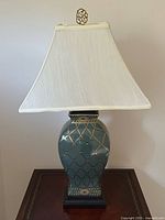 Full front view of the lamp placed on a table showing shade, finial, and ceramic body