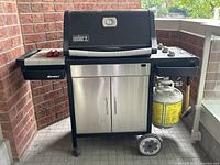 Front view of Weber Spirit propane grill showing black hood with Weber logo, two side shelves, stainless steel cabinet and attached propane tank