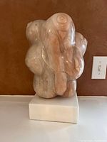 View of abstract marble sculpture signed by 'Roni', showing rounded breast forms from a front angle on a marble base against a brown background.