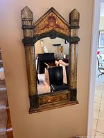 Decorative wall mirror with gold base and hand-painted finish, shown against a wall with furniture seen reflecting in the mirror.