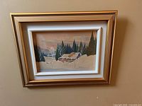 Framed watercolour painting by Graham Norwell showing a snowy cabin in a forest setting, signed on the bottom right corner.
